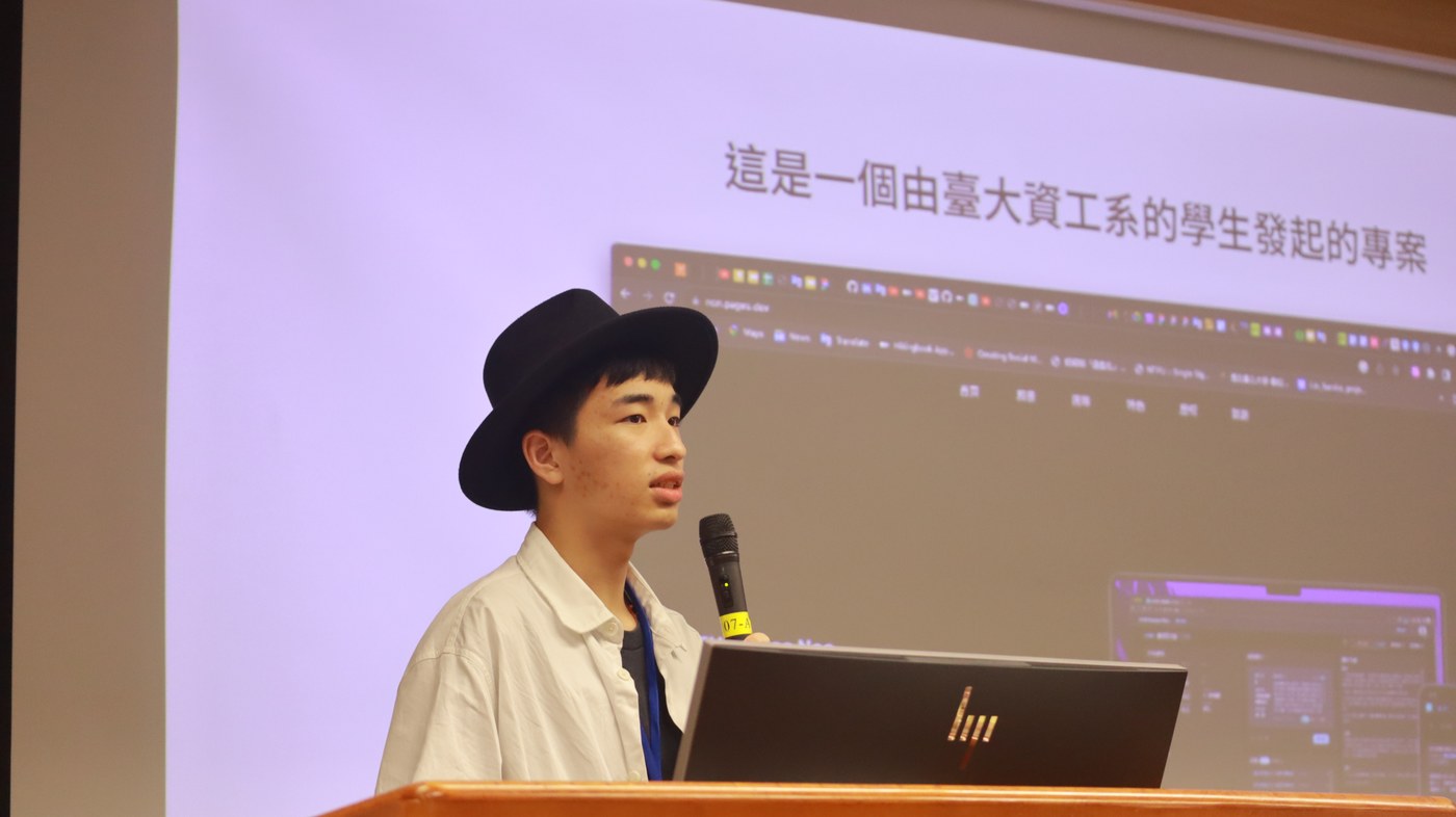 Tofus Wang, wearing a black hat and white shirt, speaks into a microphone behind a lectern. The projection introduces a project initiated by NTU computer science students.